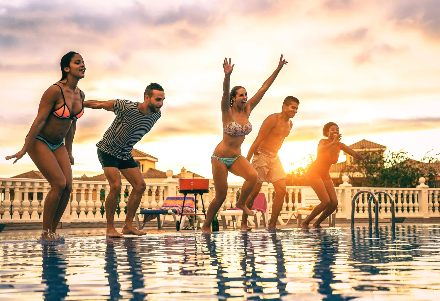 Group of happy friends jumping in the pool at sunset - Young people having fun making party in exclusive resort tropical in vacation - Concept of friendship, holidays and youth lifestyle<br />